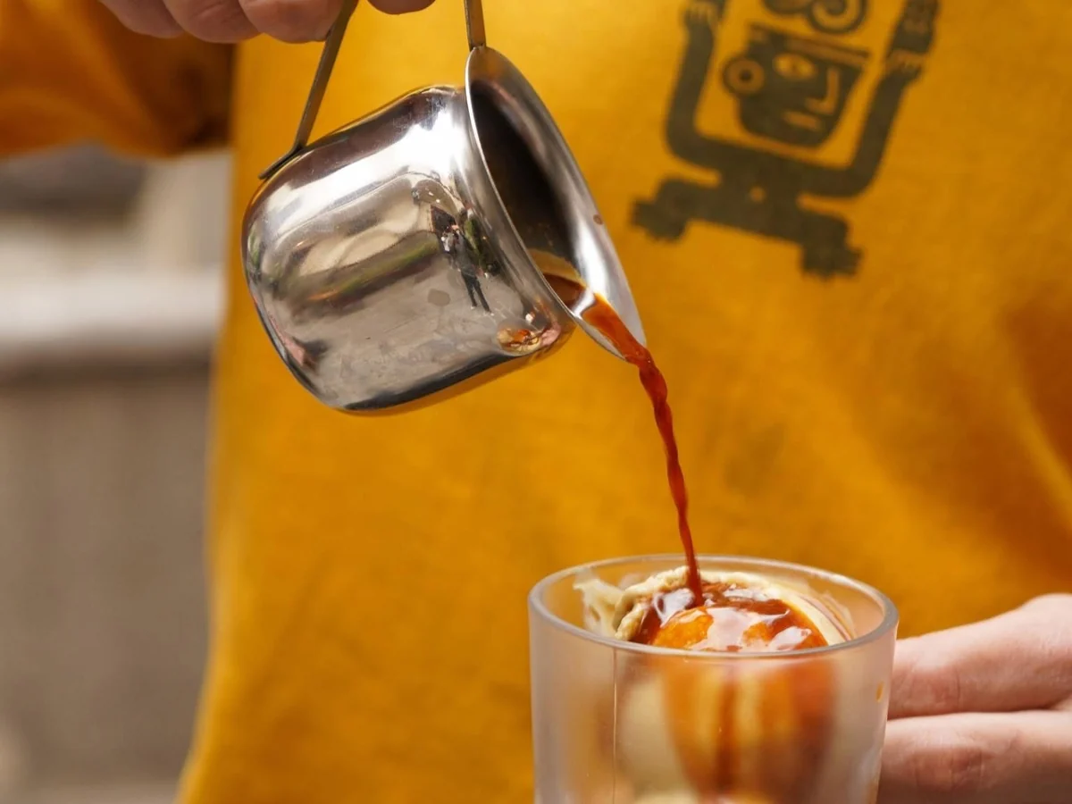 Espresso servido desde una jarrita de acero sobre helado de vainilla, frente a una playera con la figura de Esencia y Raíz
