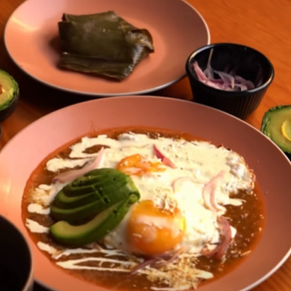 Huevos estrellados en salsa roja con aguacate y cebolla morada, y un tamal envuelto en hoja al fondo